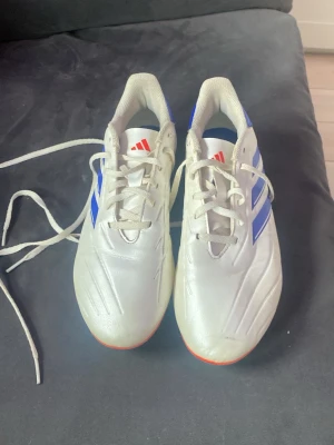 Vita Adidas fotbollsskor med blå ränder - Säljer ett par vita fotbollsskor från Adidas med klassiska blå ränder på sidorna och röda detaljer på plösen. Skorna har snörning och en låg profil, perfekta för snabba rörelser på planen. Yttermaterialet är syntetiskt och designen är sportig och modern.