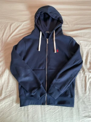 Mörkblå hoodie från Polo Ralph Lauren - Mörkblå hoodie med dragkedja från Polo Ralph Lauren. Klassisk design med vit dragsko i huvan, två fickor framtill och röd broderad logga på bröstet. Mjuk insida i fleece och ribbade muddar vid ärmar och nederkant. Perfekt för en avslappnad stil.