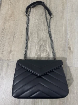 Svart quiltad handväska med kedjerem - Snygg svart handväska i quiltad design med V-format mönster. Väskan har en lång silverfärgad kedjerem med axelparti i läderimitation. Perfekt för dig som gillar stilrena accessoarer med en modern touch.