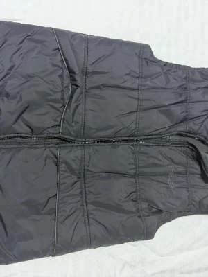 Svart dunväst med dragkedja - Snygg svart dunväst med quiltad design och dragkedja framtill. Västen har ståkrage och diskreta sömdetaljer som ger en sportig look. Perfekt att slänga över en hoodie eller tröja för extra värme. Materialet är glansigt och känns lätt men ändå isolerande.