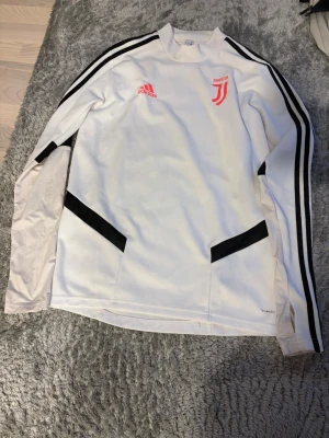 Vit Juventus träningströja Adidas - Snygg vit långärmad träningströja från Adidas med Juventus-logga och svarta detaljer. Tröjan har svarta ränder längs ärmarna och röda Adidas- och Juventusmärken på bröstet. Tillverkad i funktionsmaterial som andas, perfekt för träning.