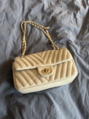 Handväska från Gina Tricot - Snygg beige axelväska i quiltat mönster med guldfärgad kedjerem och vridlås framtill från Gina Tricot. Väskan har en klassisk rektangulär form och är tillverkad i konstläder. Gott skick med lite färgskillnad på spännet. Se sista bild för liten skavank på axelremmen. Inget som syns märkbart eller stör. 
