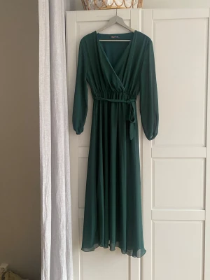 Gön sommarklänning - Elegant grön klänning med v-ringning. Klänningen har långa ärmar med lätt puff och ett knytband i midjan för en smickrande siluett. Tillverkad i ett tunt, flytande polyestertyg som ger ett mjukt fall. Klänningen är märkt 3 XL men passar mig som är L/ XL.