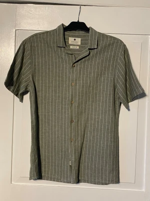 Grön randig linneskjorta  - Randig skjorta i linnmix från anerkjendt. Trevlig struktur med schyssta detaljer så som träknappar samt en diskret logopatch längst ner i knappöpningen. Relaxed fit enligt lappen men sitter mer regular/slim. Sitter M-S. Säljer billigt då jag desperat behöver plats i garderoben 😅