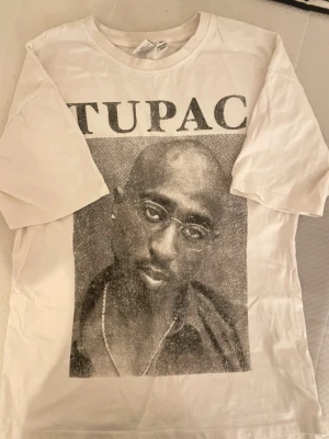 Vit Tupac t-shirt med tryck - Säljer en vit t-shirt  med stort svartvitt Tupac-tryck på framsidan. T-shirten har rund hals och korta ärmar, perfekt för dig som gillar streetwear och vill ha en ikonisk look.