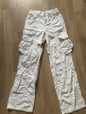 Vita cargobyxor från H&M Divided - Säljer ett par vita cargobyxor från H&M Divided med bred resår i midjan och stora fickor på sidorna. Byxorna har en avslappnad passform och är tillverkade i ett lätt bomullsmaterial. Perfekta för en chill och trendig look.