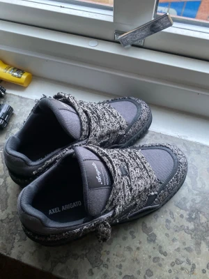 Gråa sneakers Axel Arigato - Säljer ett par gråa sneakers från Axel Arigato. Skorna har chunky sula, breda mönstrade skosnören och en mix av textil och mockaliknande material. Snygg och modern design som sticker ut med sin unika struktur och färgkombination. 