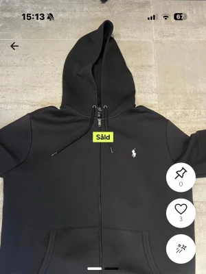 Svart zip hoodie från Polo Ralph Lauren - Svart hoodie med dragkedja från Polo Ralph Lauren. Klassisk design med vit broderad logga på bröstet, huva med dragsko och kängurufickor framtill. Tillverkad i mjukt bomullsmaterial som är skönt att bära.