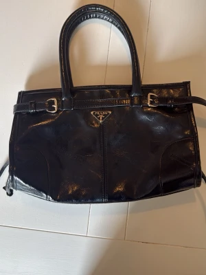 Svart handväska i skinn från Prada - Snygg svart handväska i glansigt skinn med silverfärgade detaljer och klassisk trekantig logga framtill. Väskan har dubbla handtag och dekorativa spännen. INTE ÄKTA 