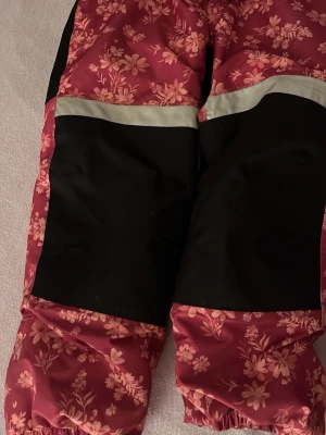 Blommiga täckbyxor i rosa och svart - Täckbyxor i rosa med blommönster och svarta förstärkningar på knäna. Reflexdetaljer och resår vid benslut. Perfekta för kalla dagar och utomhusaktiviteter. Materialet är vattenavvisande och vindtätt för extra komfort.