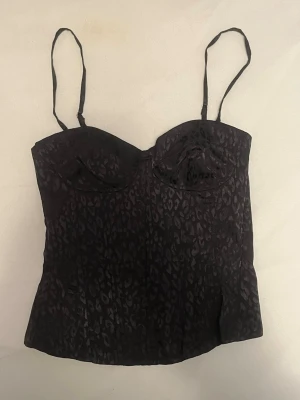 Svart leopardmönstrad korsettopp - Snygg svart korsettopp från Bershka (oanvänd) med leopardmönster och tunna justerbara axelband. Toppen har en figurnära passform och lätt vadderade kupor för extra stöd. Perfekt för att lyfta din outfit med en edgy touch.