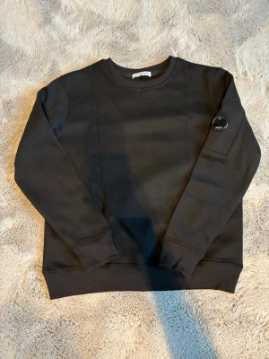 Svart sweatshirt från C.P. Company - Säljer en svart sweatshirt från C.P. Company med deras ikoniska linsdetalj på ärmen. Tröjan har rund halsringning, ribbade muddar och är tillverkad i mjukt bomullsmaterial. Perfekt för en clean och stilren look.