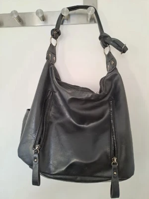 Svart axelremsväska med dragkedjor - Cool svart handväska i mjukt konstläder med två långa dragkedjor framtill och justerbar axelrem. Väskan har en slouchy form och silverfärgade metalldetaljer. Perfekt för dig som gillar en stilren men edgy look. Den är väl använd och de syns tyvärr lite på en av dragkedjorna men den är fortfarande väldigt fin och användbar. 