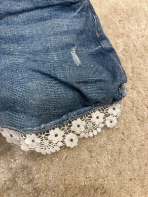 Blå jeansshorts med spetskant - Snygga blå jeansshorts med råa kanter och vit spetsdetalj längst ner. Shortsen har klassisk femficksdesign och slitna detaljer framtill för en trendig look. Perfekta för varma dagar och ger en söt touch med spetsen.