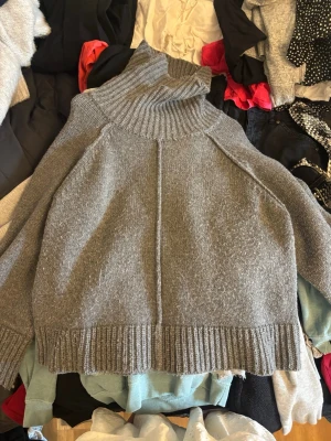 Grå stickad polotröja - Mysig grå stickad polotröja med bred ribbad kant vid ärmslut och nederkant. Tröjan har en hög polokrage och avslappnad passform, perfekt för lager på lager. Materialet ser ut att vara mjukt och värmande, troligen ull- eller ullmix.