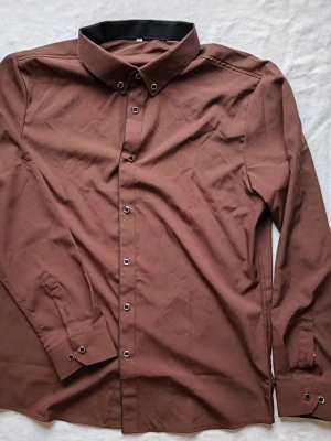 Brun skjorta med svarta detaljer - Stilren brun skjorta med svarta detaljer i kragen och svarta knappar. Skjortan har lång ärm och klassisk krage, perfekt för en clean och modern look. Passar dig som gillar minimalistisk stil.