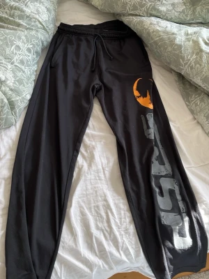 Svarta gymbyxor från GASP - Säljer ett par svarta mjukisbyxor från GASP med stort tryck i vitt och orange längs ena benet. Byxorna har dragsko i midjan och är tillverkade i ett mjukt och stretchigt material. Perfekta för chill eller gymmet.