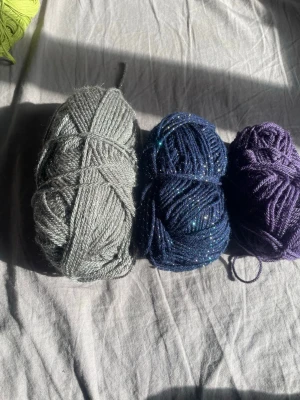 Tre nystan garn i olika färger - Säljer tre nystan garn: ett ljusgrått, ett mörkblått med glittereffekt och ett lila. Perfekt för stickning eller virkning av egna projekt. Garnet är mjukt och passar till både accessoarer och dekorationer.