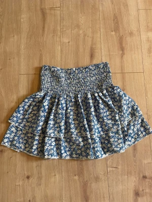 Blommig blå kortkjol med volang - Supersöt kortkjol i blått med vitt blommönster och bred smock i midjan. Kjolen har volangdetaljer och är perfekt för en somrig look. Materialet känns lätt och luftigt.