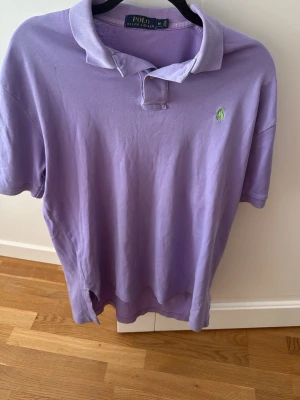   Polo Ralph Lauren piké - Säljer en lila pikétröja från Polo Ralph Lauren i storlek M. Tröjan har klassisk krage, korta ärmar och den ikoniska gröna loggan broderad på bröstet. Tillverkad i mjuk bomull som är skön att bära. Perfekt för en avslappnad och stilren look.