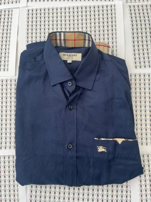 Mörkblå skjorta från Burberry - Stilren mörkblå skjorta från Burberry med klassiskt rutigt mönster på insidan av kragen och manschetterna. Skjortan har en bröstficka med Burberry-logga och knappstängning framtill. Perfekt för dig som gillar exklusiva detaljer och tidlös design.