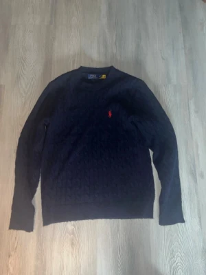  Ralph Lauren tröja - Kabelstickad mörkblå tröja från Polo Ralph Lauren med rund halsringning och den klassiska röda logotypen broderad på bröstet. Storlek S