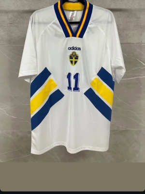 Svensk landslagströja Adidas Brolin - Vit svensk landslagströja från Adidas med blå och gula detaljer, nummer 11 och Brolin på ryggen. Tröjan har korta ärmar, V-ringad krage med blå och gula ränder samt broderad svensk landslagssköld på bröstet. Perfekt för fotbollsfans!