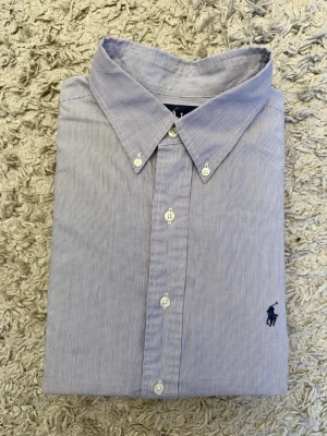 Randig skjorta från Ralph Lauren - Klassisk blå- och vitrandig skjorta från Ralph Lauren med button-down krage och broderad logga på bröstet. Skjortan har långa ärmar och knäppning framtill. Tillverkad i mjuk bomull för en bekväm känsla. Sitter som L/XL