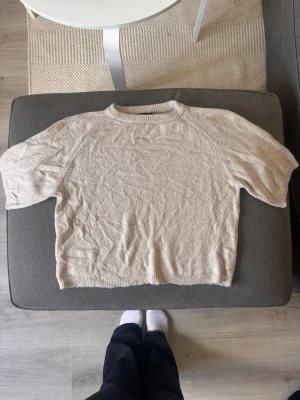 Beige stickad tröja från 157 - Beige stickad tröja från 157 med kortare ärmar och rund halsringning. Tröjan har en avslappnad passform och ribbstickade detaljer vid hals, ärmslut och nederkant. Perfekt för lager-på-lager och enkel att matcha med olika outfits.