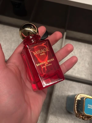 Mirage Paris Sandal Musk parfym - En lyxig parfym från Mirage Paris med doften Sandal Musk. Flaskan är fyrkantig i rött glas med guldfärgat klotformat lock och elegant guldtext. Perfekt för dig som gillar exklusiva och djupa dofter.   KAN OCKSÅ BYTAS!