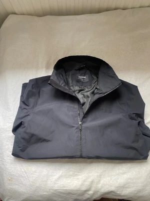 Svart vindjacka från Everest - Snygg svart vindjacka från Everest med dragkedja framtill och hög krage. Jackan har två framfickor och justerbara muddar med kardborre. Tillverkad i ett lätt och slitstarkt material som står emot vind. Perfekt för dig som vill ha en stilren och funktionell jacka. 