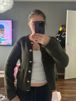 Mörkgrön ribbad kofta med fickor - Snygg mörkgrön ribbstickad kofta med rund halsringning och guldknappar. Koftan har två framfickor och långa ärmar. Perfekt att slänga över en topp för en chill och trendig look.