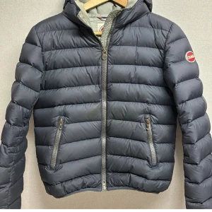 Mörkblå dunjacka från Colmar - Snygg mörkblå dunjacka från Colmar med huva och två dragkedjeförsedda fickor. Jackan har quiltad design och Colmar-logga på ärmen. Perfekt för kalla vinterdagar och tillverkad i lätt, värmande material.