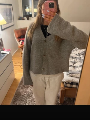 Grå kofta med knappar - Mysig grå kofta i  modell med v-ringning och stora knappar framtill. Koftan har långa ärmar och är något kortare i modellen, vilket ger en avslappnad look. Perfekt att slänga över en t-shirt eller linne för en chill stil.