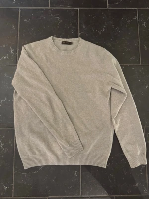 Beige stickad tröja från Oscar Jacobson - Snygg beige stickad tröja från Oscar Jacobson med rund halsringning och ribbade muddar vid ärmslut och nederkant. Tröjan har en enkel och stilren design som passar perfekt till många olika outfits. Materialet ser mjukt och bekvämt ut.