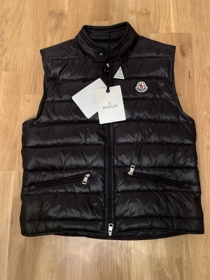 Svart dunväst från Moncler - Snygg svart dunväst från Moncler, modell Longue Saison. Västen har en glansig finish och är lätt vadderad med dun för extra värme. Perfekt för lager-på-lager och ger en stilren look. Klassisk design med Moncler-logga på insidan. NFC finns, priset kan diskuteras. skicka dm vid flera bilder.