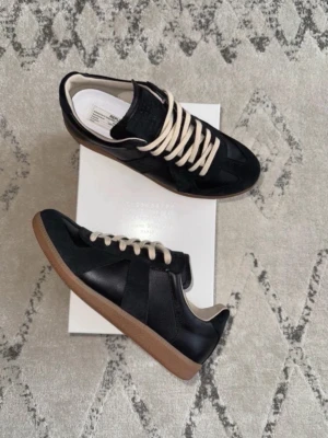  Maison Margiela gats - Snygga svarta sneakers från Maison Margiela med beige sula och vita skosnören. Skorna har en klassisk låg silhuett och är tillverkade i en mix av läder och mocka. Perfekta för dig som gillar stilrena och tidlösa sneakers med en lyxig känsla.