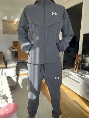 Grått loungewear-set från Under Armour - Snyggt grått loungewear-set från Under Armour med matchande hoodie och byxor. Hoodien har dragkedja, justerbar huva och svarta loggor på bröstet och byxan. Byxorna har resår i midjan och dragkedjeförsedda fickor. Perfekt för chill dagar hemma.