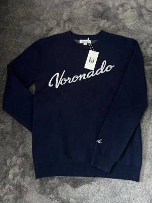 Mörkblå stickad tröja Voronado Atelier - Mörkblå stickad tröja från Voronado Atelier med vit broderad logga över bröstet. Tröjan har rund halsringning och långa ärmar. Perfekt till jeans för en clean och stilren look. Materialet känns mjukt och bekvämt.