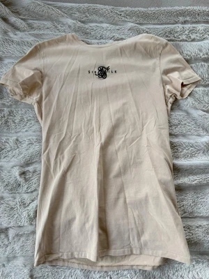 Beige t-shirt från SikSilk - Snygg beige t-shirt från SikSilk med diskret logga i svart på bröstet. Klassisk passform med rund hals och korta ärmar. Tillverkad i mjuk bomull som känns skön mot huden. Perfekt till jeans eller shorts för en clean look.
