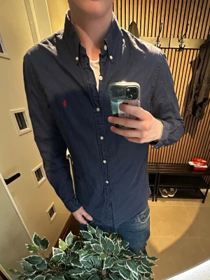 Mörkblå skjorta från Ralph Lauren - Klassisk mörkblå skjorta från Ralph Lauren med button-down krage och röda logotypen broderad på bröstet. Skjortan har långa ärmar, vita knappar och är tillverkad i bomull. Perfekt till jeans för en clean och stilren look.