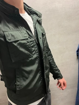 Mörkgrön vindjacka Massimo Dutti - Stilren mörkgrön vindjacka från Massimo Dutti med två stora bröstfickor och dragkedja framtill. Jackan har hög krage, knappdetaljer och är tillverkad i ett lätt, glansigt material som passar perfekt för blåsiga dagar.