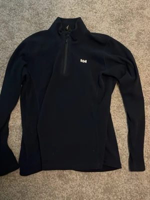 Svart fleece tröja från Helly Hansen - Mjuk och skön svart fleece tröja från Helly Hansen med halv dragkedja framtill och diskret logga på bröstet. Perfekt för lager-på-lager och har en sportig, enkel design med lång ärm och hög krage.