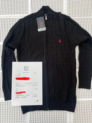 Svart kabelstickad kofta Polo Ralph Lauren - Svart kabelstickad kofta från Polo Ralph Lauren med dragkedja och röd broderad logga på bröstet. Koftan har ribbade muddar och klassisk krage. Perfekt för lager-på-lager och snygg till jeans.