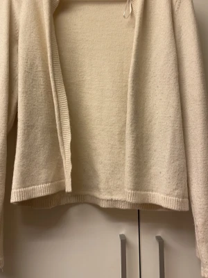 Beige stickad öppen kofta - Mysig beige stickad kofta med öppen framsida och långa ärmar. Koftan har ribbade muddar vid ärmslut och nederkant, vilket ger en avslappnad och enkel look. Perfekt att slänga över en t-shirt eller topp för extra värme.