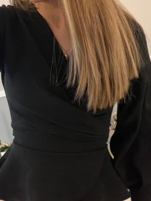 Svart omlottblus med peplum - Trendig svart omlottblus med peplumdetalj i midjan och långa ärmar. Blusen har en smickrande v-ringning och markerad midja som ger en snygg siluett. Perfekt att styla med både jeans och kjol. Har även knyte bak, skriv för bild bak💓