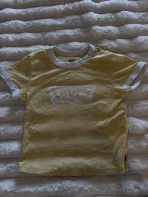 Gul Levi's t-shirt med vit logga - Snygg gul t-shirt från Levi's med vit logga på bröstet och vita detaljer vid krage och ärmslut. Klassisk passform och tillverkad i mjuk bomull, perfekt för en avslappnad stil. 30cm i midjan.