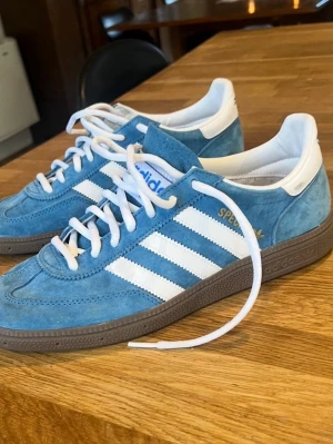 Adidas Spezial blå sneakers mocka - Snygga Adidas Spezial sneakers i blå mocka med vita detaljer och klassiska tre ränder på sidan. Skorna har vit hälkappa, vita skosnören och en mörkbrun yttersula. Perfekta för dig som gillar retrostil och streetwear.