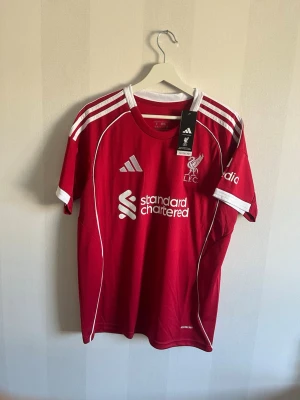Liverpool matchtröja Adidas röd - Röd Liverpool matchtröja från Adidas med vita detaljer och tryck. Tröjan har klubbmärke, sponsorlogga och nummer 9 med namnet ISAK på ryggen. Korta ärmar, rund hals och tillverkad i lätt funktionsmaterial. Perfekt för fotbollsträning eller support.