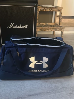 Blå träningsväska Under Armour - Snygg marinblå träningsväska från Under Armour med stort vitt logotryck på framsidan. Väskan har justerbar axelrem, bärhandtag och flera dragkedjefack. Tillverkad i slitstarkt syntetmaterial och har en rymlig, rektangulär form. Aldrig använt gillade inte den